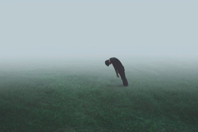 A moody photograph showing the silhouette of a person bent over looking at the ground in a grassy field. The scene is heavily obscured by thick, dense fog, creating a mysterious and isolated atmosphere with a cool, blueish-green color palette.