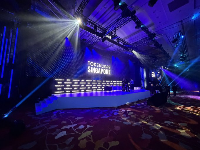 A stage setup at TOKEN2049 SINGAPORE with blue lighting, spotlights, and event branding visible on screens and panels.