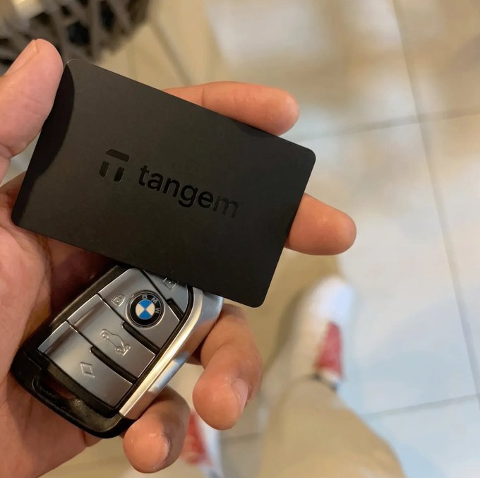 A hand holding a black Tangem crypto hardware wallet card above a BMW car key fob, with a tiled floor and the person's white sneakers visible in the background.