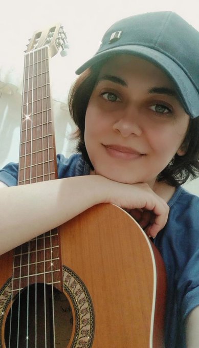 A selfie of a person wearing a green cap, smiling while leaning on a classical acoustic guitar.