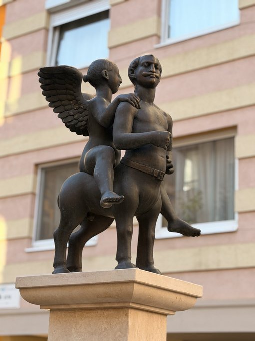 A bronze statue depicting a centaur-like creature with a human upper body carrying a smaller winged humanoid figure on its back, displayed on a stone pedestal in front of a building with pink and yellow walls.
