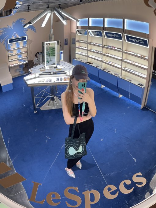 A woman takes a mirror selfie in a LeSpecs eyewear store while wearing Hyperliquid merchandise, including a black "Hype" cap and a beaded lanyard, with a brand-logo tote bag on her shoulder. The "LeSpecs" branding is prominently displayed on the mirror, matching the post's mention of showcasing new Hyperliquid merch.