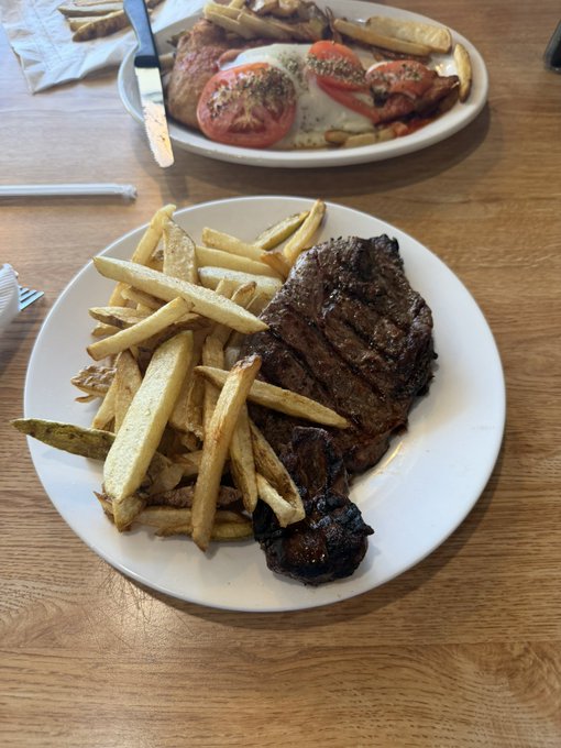 Two plates of food sit on a wooden table, one featuring a grilled steak with french fries and the other with tomatoes, eggs, and bacon.