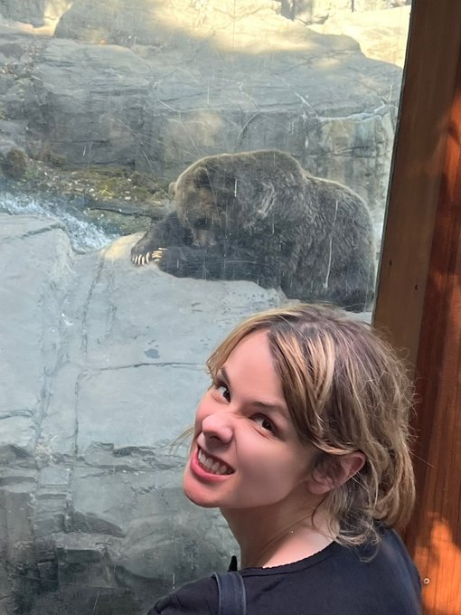A young girl with a bright smile poses in front of a glass enclosure where a large brown bear is visible behind her, looking down at the ground. The playful caption "$dasha" humorously suggests that even the bear behind the glass is afraid of the confident child in the foreground.