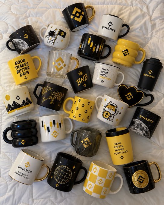 A collection of Binance-branded mugs in black and yellow featuring various designs—including astronaut graphics, world maps, candlestick charts, and slogans like "GOOD TRADES BETTER DAYS," "BUILD TRADE HOLD REPEAT," "BNB," and "SAME COFFEE HIGHER PORTFOLIO"—arranged on a white sheet to accompany a "GM Coffee first" morning post asking followers which mug they'd grab.