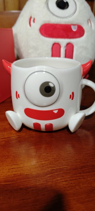A novelty ceramic mug designed as a one-eyed cartoon monster with horns, red mouth, and small arms, sitting on a wooden table in front of a matching plush toy.