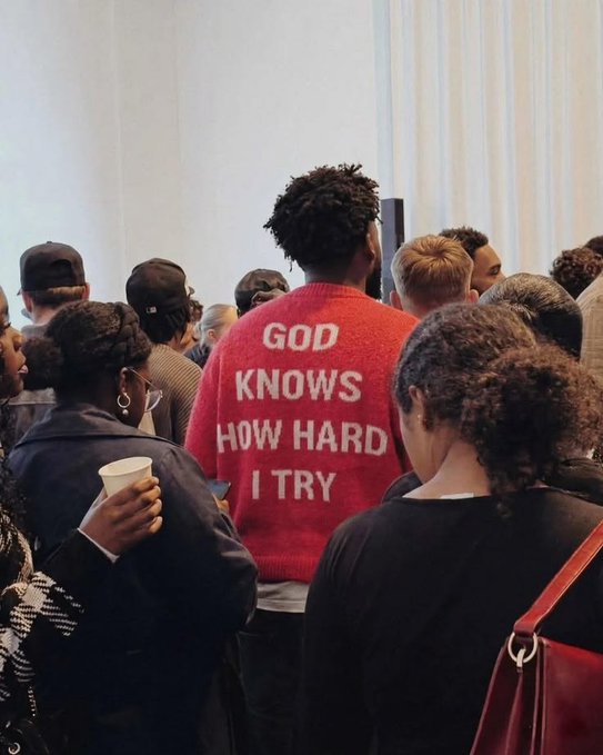 A person viewed from behind wearing a red sweater that reads 'GOD KNOWS HOW HARD I TRY' stands among a crowd.