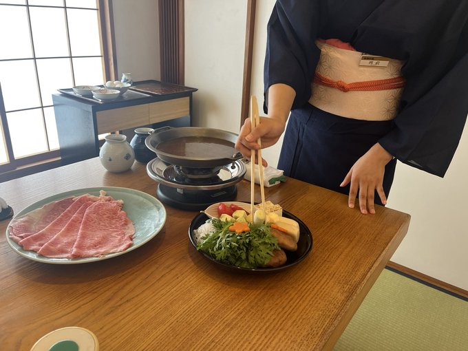 A server in a traditional kimono (with a nametag reading "西谷") serves a beautifully arranged sukiyaki meal on a low table in a tatami room, featuring thinly sliced marbled beef, fresh vegetables, tofu, and a simmering pot of broth, perfectly capturing the authentic Japanese dining experience that made the traveler fall in love with Japan over Dubai.