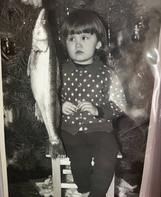 A vintage black-and-white photo shows a young child in a polka-dot shirt and dark tights holding up a large fish next to a decorated Christmas tree, humorously presented as the "boss" of the account with an early love for fishing.