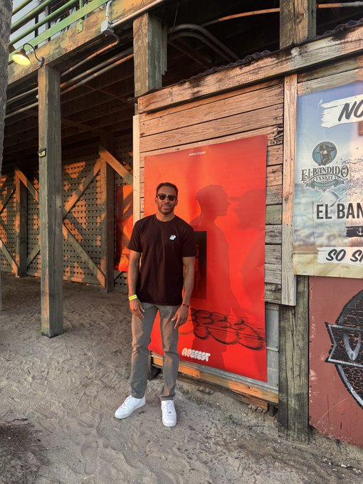 A man poses in front of a red poster near an "El Bandido" sign at what appears to be the ApeFest event. The rustic wooden beachside setting suggests the festival's casual, immersive atmosphere.