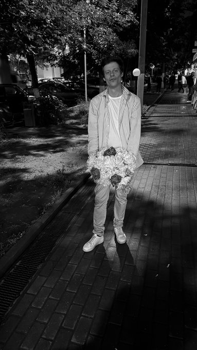 A black and white photograph of a smiling young man standing on a brick-paved sidewalk at night, holding a bouquet of flowers while wearing a striped shirt.
