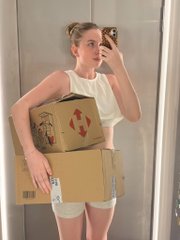 A mirror selfie of a woman in a white tank top and shorts holding stacked cardboard moving boxes, including one with a red label.