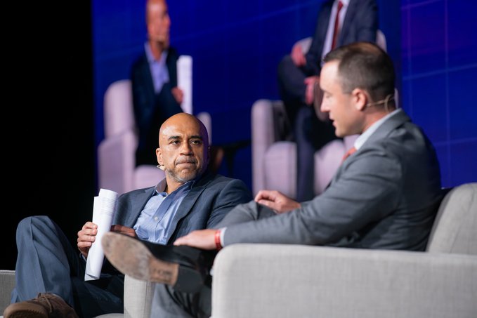 Two men in suits sit in armchairs on a blue-lit stage, appearing to speak during a panel discussion or interview.