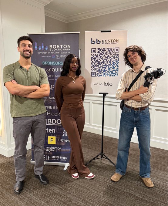 Three people pose in front of banners for 'BOSTON BLOCKCHAIN' which lists the date September 8th-10th, 2025 in Quincy, MA, along with sponsor logos and a QR code. The group includes a man in a green henley, a woman in a brown outfit, and a man holding a camera wearing a striped shirt.