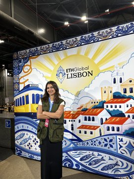 A woman with glasses and an olive jacket stands with arms crossed in front of a mural reading 'ITMGlobal LISBON' depicting Portuguese-style buildings.