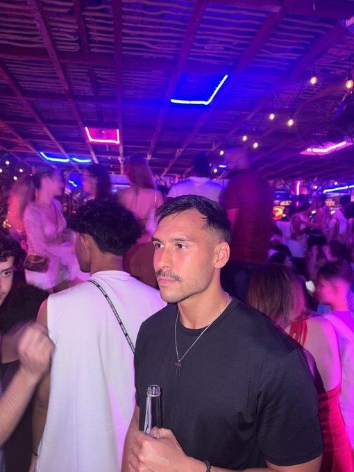 The image shows a man holding a beer bottle, surrounded by other people at an event with purple lights overhead. There are also several people standing around the room, some of whom appear to be engaged in conversation or enjoying themselves. The scene suggests that this gathering is taking place during a party or social event where attendees are mingling and having a good time.