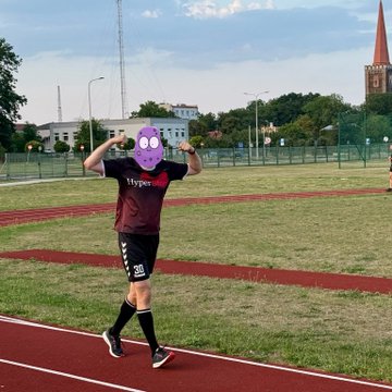 A person running on a track at a stadium, wearing a maroon shirt and black shorts, with their face obscured by a purple skull emoji, flexing their arms in celebration.
