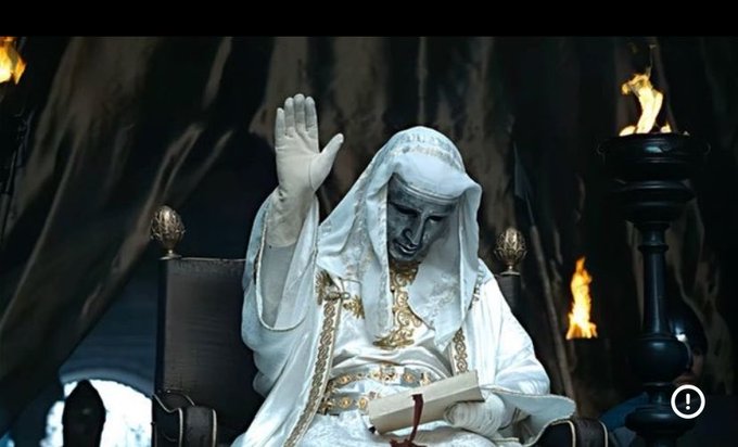 A dramatic scene featuring a masked, pale figure in elaborate white and gold robes sitting on a throne, holding a book and raising a hand, with flaming torches in the dark background.
