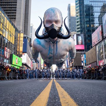 Giant inflatable mascot balloon in an urban city street setting, with the inflatable figure wearing a gas mask-like face covering and surrounded by crowds of people.