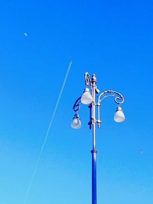Blue vintage street lamp with three white globes against a clear deep blue sky, with the moon visible in the upper left and a jet's contrail streaking across the frame.