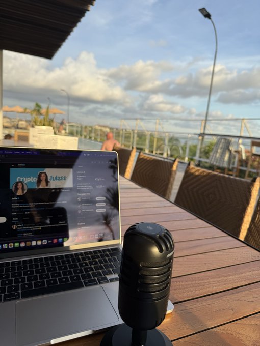 A laptop displaying what appears to be a social media feed with a post titled "Crypto Juizz" sits next to a black microphone on an outdoor wooden deck overlooking a scenic waterfront, fitting the "apes in" vibe of recording crypto or meme commentary content.