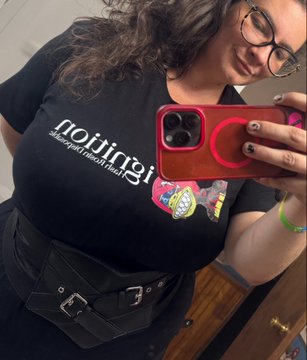 A mirror selfie shows a person with dark curly hair and glasses wearing a black 'notting hill' t-shirt featuring a colorful graphic, holding a phone with a red case and a red circular pop socket.