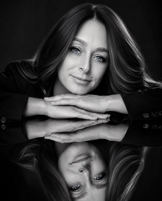 A black-and-white portrait of a woman with long wavy hair resting her chin on her hands on a reflective surface, with her reflection visible below.