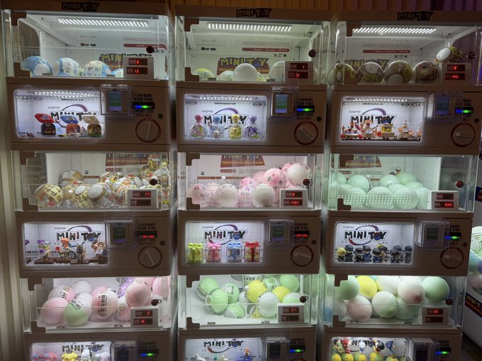 A wall of illuminated gachapon capsule toy vending machines labeled 'MINI TOY', filled with various colorful capsule toys in clear balls.