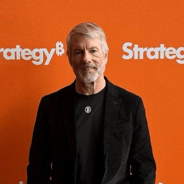 An older man with gray hair and beard, wearing a black jacket and t-shirt, poses in front of an orange backdrop with the text 'Strategy' visible.