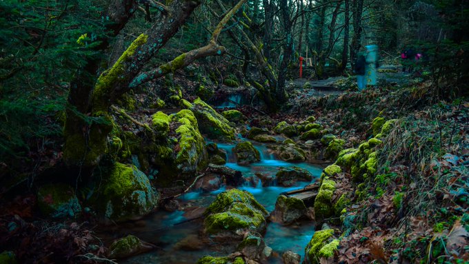 A mossy forest stream flowing over rocks, captured with a long exposure effect creating smooth blue water, with figures faintly visible in the background.