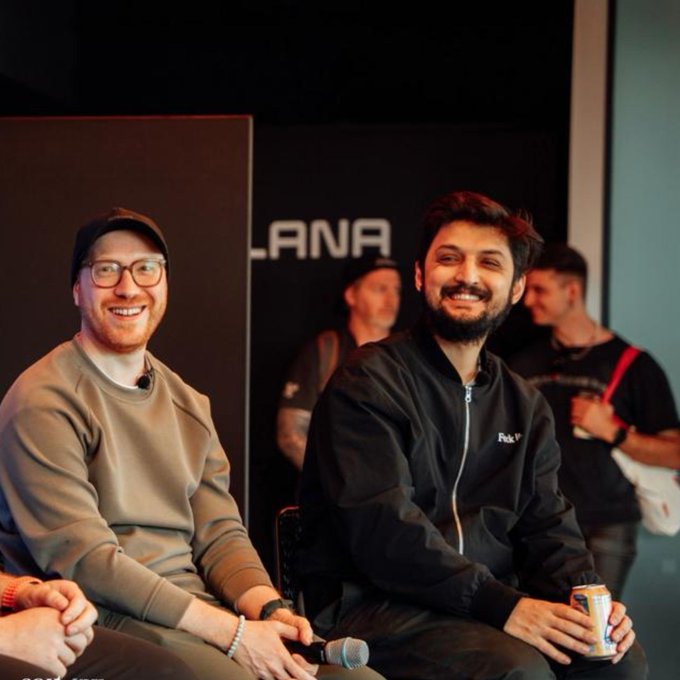 Two smiling men are seated together at an event with 'LANA' visible on the backdrop behind them; the man on the right holds a beverage can and wears a jacket with 'Park N°' on it.