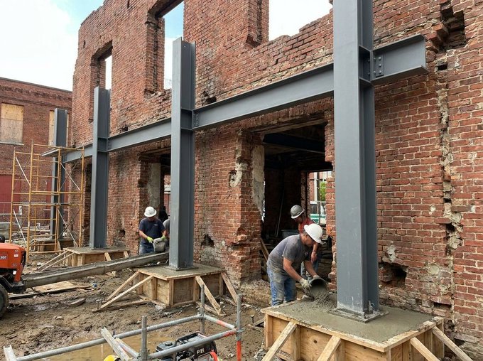 Construction workers in hard hats are pouring concrete into wooden forms at the base of new steel support columns inside a dilapidated red brick building.
