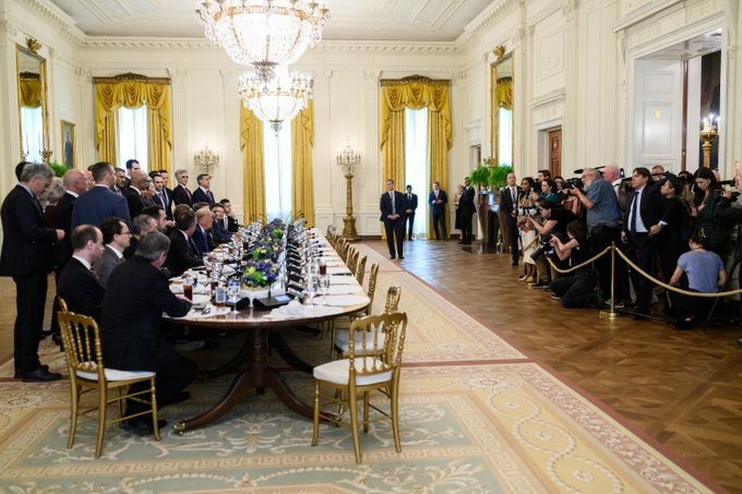 A formal gathering in an ornate room with crystal chandeliers, where officials in suits sit at a long table while photographers and observers watch.