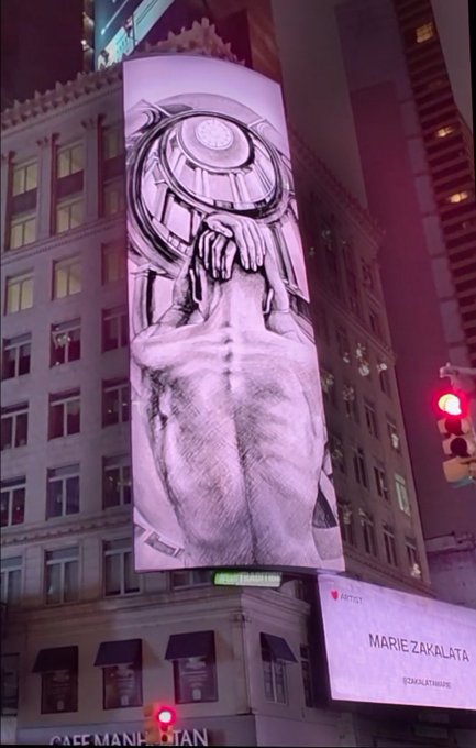A large illuminated billboard on a city building displaying a pencil-sketch artwork of a woman's body with a camera for a head. Below it, another billboard reads 'MARIE ZAKALATA' and 'CAFE MANEKIATAN' is visible at street level.
