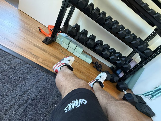 A first-person view of someone wearing Adidas sneakers and black "Quirkies" shorts sitting in front of a rack of dumbbells, likely resting between sets during a fitness session while repping their QuirkiesNFT gear.