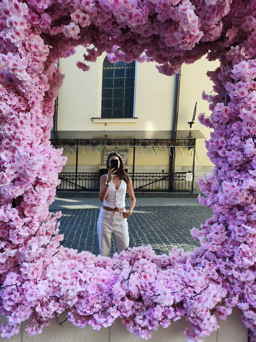 A mirror framed with pink cherry blossoms reflects a person in white clothing taking a selfie with their phone in an outdoor courtyard setting.