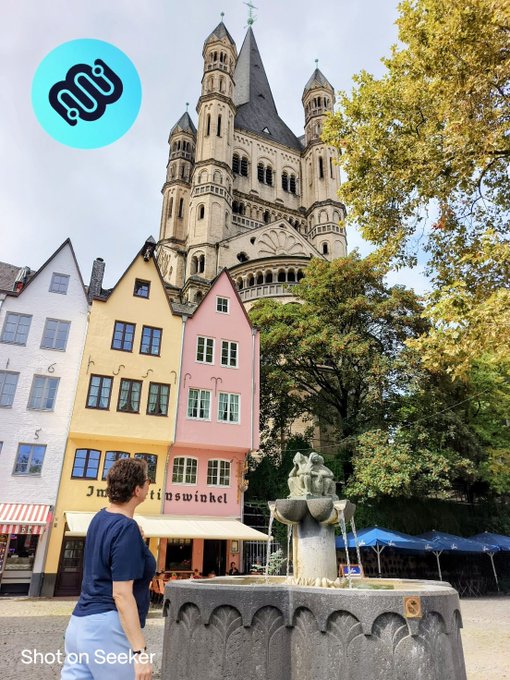 A photo taken in Cologne (Köln), Germany, showing colorful traditional houses and a fountain in the foreground with the Great St. Martin Church in the background, with a person walking nearby.