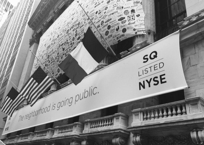 A black and white photo of the New York Stock Exchange building with a banner reading 'SQ LISTED NYSE' and 'The neighborhood is going public.' along with American flags.