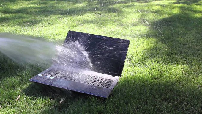 An open dark laptop sits on green grass while a stream of water sprays onto its keyboard, splashing off the screen.