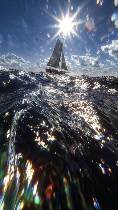 A sailboat on a sunlit ocean with dramatic lighting, featuring the sun shining brightly above the boat and sparkling light reflecting across the wavy water.