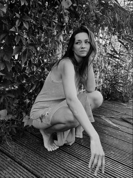 A black-and-white photograph of a woman crouching barefoot on a wooden deck, wearing a light-colored slip dress, with foliage behind her.