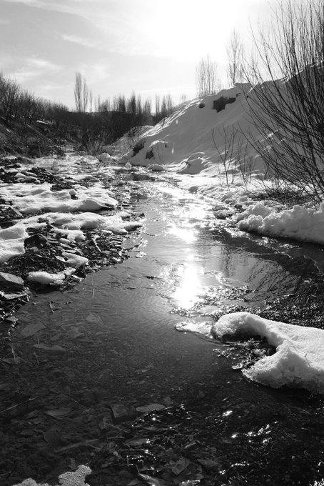 A black-and-white photograph captures a partially frozen stream winding through a snowy landscape, with sunlight glinting off the open water and ice chunks scattered along its banks. The stark, serene winter scene evokes the contemplative mood of the artwork "You were the light," a 4/10 edition Tezos NFT collection by @jayray7_eth being celebrated in the post.