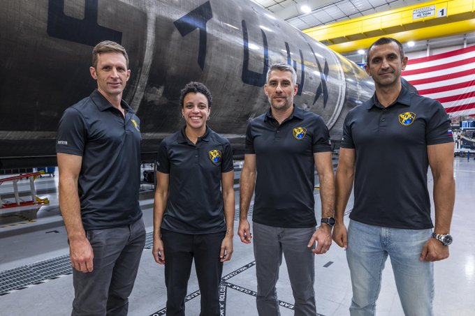 Four individuals in matching black polo shirts with 'UAE' logos stand together in an industrial setting, with a 'LINX' spacecraft component and an American flag visible behind them.