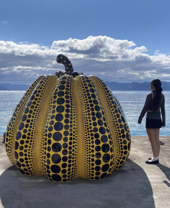A large yellow pumpkin sculpture covered in black polka dots of varying sizes sits on a concrete pier by the sea, with a person standing next to it for scale.