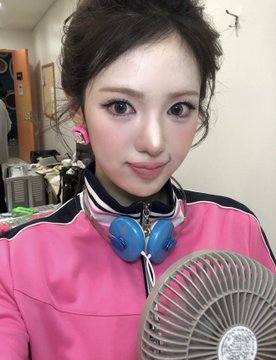 A selfie of a woman with styled hair and makeup, wearing a pink jacket with blue headphones around her neck, sitting near a fan.