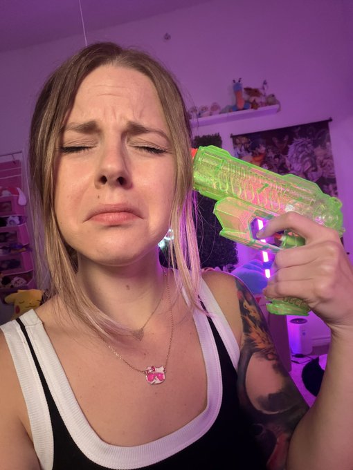 A woman with blonde hair closes her eyes with a pained expression while holding a green translucent toy water gun to her head in a purple-lit room.