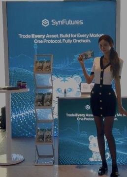 A woman stands smiling at the SynFutures booth, holding up one of the Pokémon cards given away at the event, with the company's slogan "Trade Every Asset, Build for Every Market. One Protocol. Fully Onchain." displayed on the backdrop behind her. The display rack beside her showcases the remaining promotional Pokémon cards available for visitors.