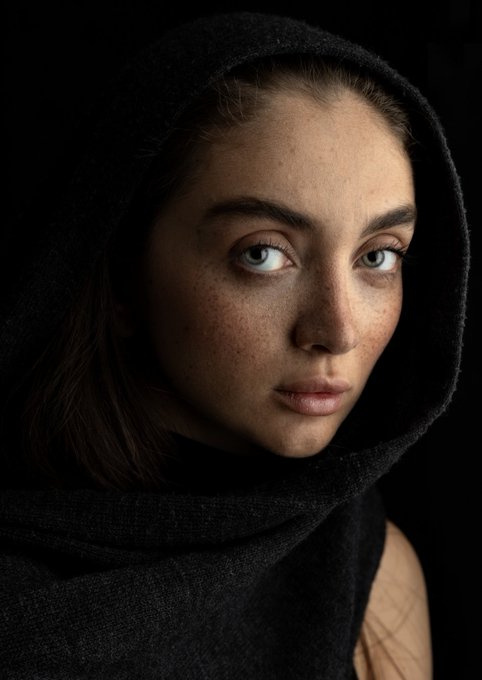 A striking studio portrait of a young woman with freckles and blue eyes, wearing a black hood and scarf against a dark background, with visible tattoos on her shoulder.