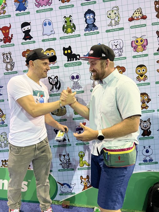 Two men shake hands in front of a wall featuring @veefriends characters. The man on the right sports a Cotopaxi fanny pack, which is a key part of the outfit mentioned in the post.
