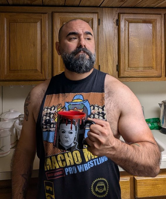 A muscular, bearded man wearing a 'Macho Man Randy Savage Pro Wrestling' tank top, holding a red coffee mug with horror character artwork on it in a kitchen.